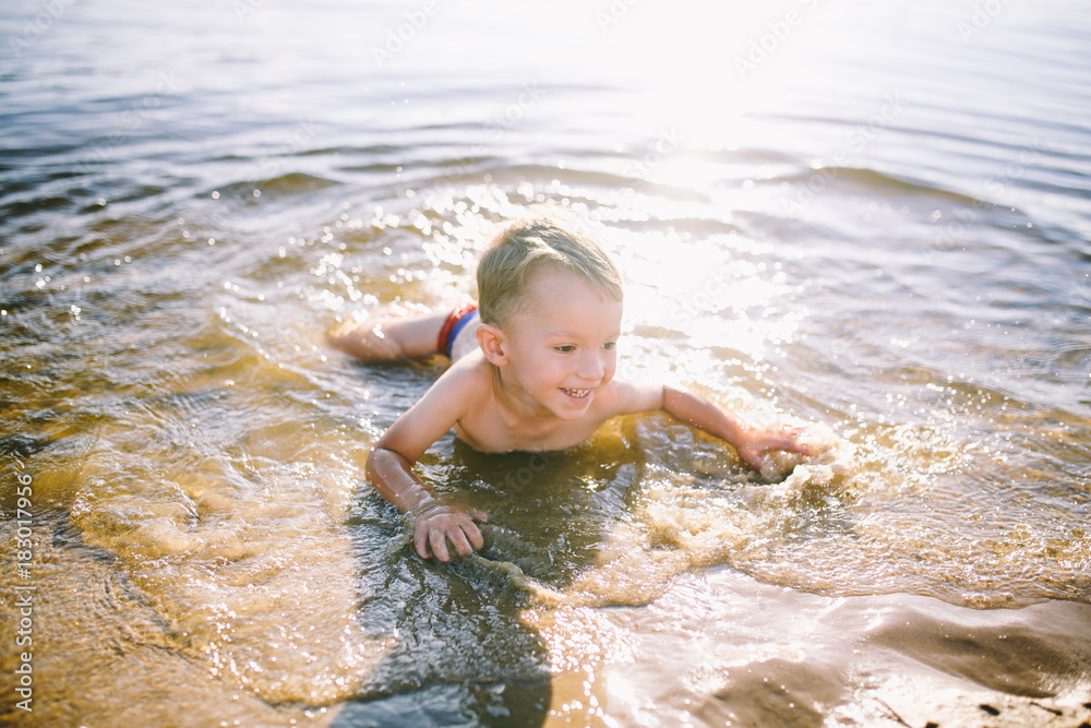 A Caucasian child of three years in red swimming trunks lies on his stomach in the water near the river bank of a sandy beach. Learns to swim with a smile A Caucasian child of three years in red swimming trunks lies on his stomach in the water near the river bank of a sandy beach. Learns to swim with a smile