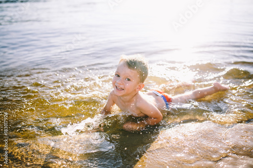 A Caucasian child of three years in red swimming trunks lies on his stomach in the water near the river bank of a sandy beach. Learns to swim with a smile