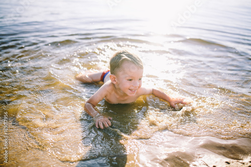 A Caucasian child of three years in red swimming trunks lies on his stomach in the water near the river bank of a sandy beach. Learns to swim with a smile