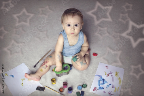 Cute boy covered in paint sitting on the floor among the items for creativity