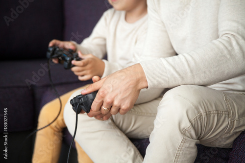 Male and kid hands holding joysticks, father and son playing video games on console together at home concept, man with little boy having fun gaming sitting on sofa in living room, close up view