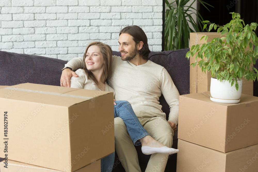 Smiling couple sitting on sofa embracing on moving in out day, married man and woman planning future after relocation into new home, family relaxing on couch in living room with cardboard boxes Smiling couple sitting on sofa embracing on moving in out day, married man and woman planning future after relocation into new home, family relaxing on couch in living room with cardboard boxes
