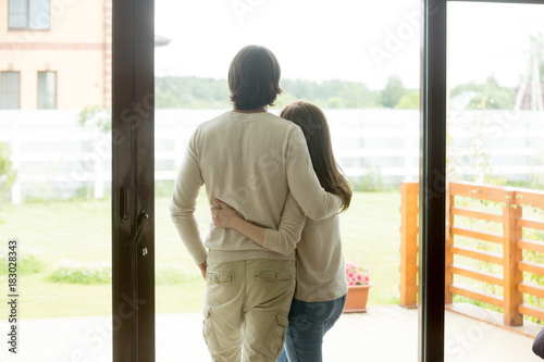 Couple standing at home, embracing looking outside through glass door of modern new house with terrace, young man and woman planning future after buying real estate, relaxing enjoying beautiful view