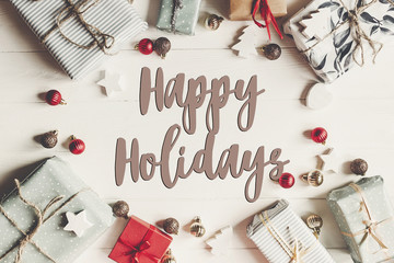 happy holidays text, seasonal greetings card sign. christmas flat lay. present boxes with ornaments on white wooden background top view, stylish wrapped gifts frame.