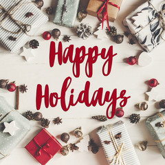happy holidays text, seasonal greetings card sign. christmas flat lay. present boxes with ornaments and pine cones anise on white wooden background top view, stylish wrapped gifts