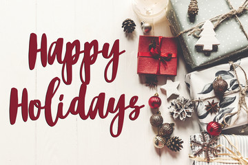 happy holidays text, seasonal greetings card sign. modern christmas wrapped presents with ornaments and cones on white wooden background top view