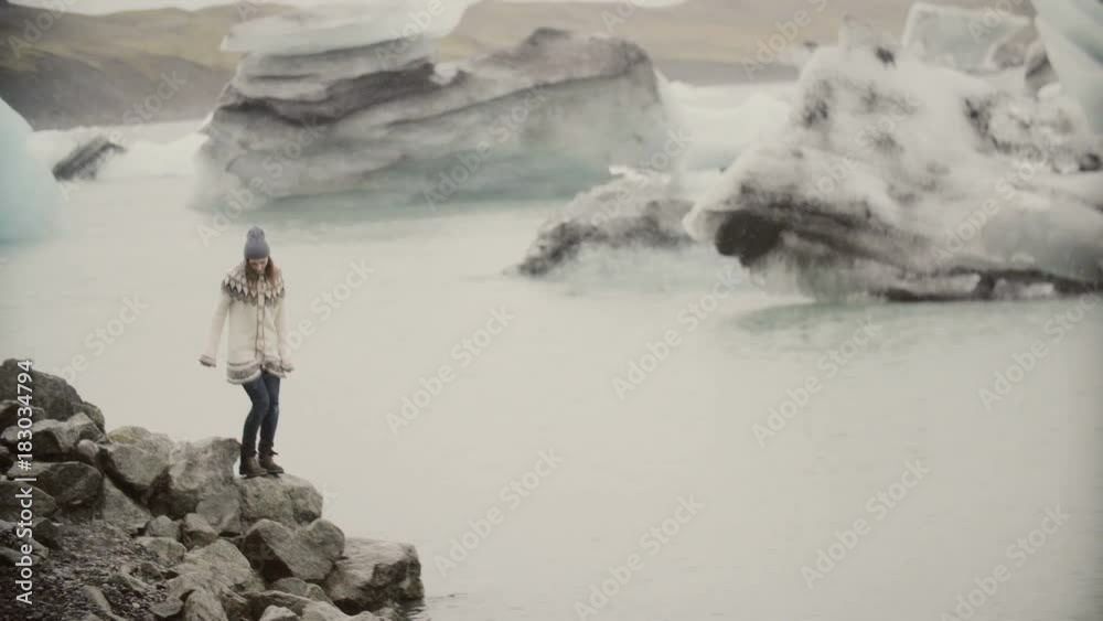 Young happy woman walking on the shore of ice lagoon. Tourist in lopapeysa exploring the sights of Iceland. Young happy woman walking on the shore of ice lagoon. Tourist in lopapeysa exploring the sights of Iceland.