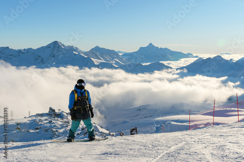 A professional snowboarder with a backpack stands on the snow high in the mountains against the background of low clouds and the Caucasus mountains during the setting sun