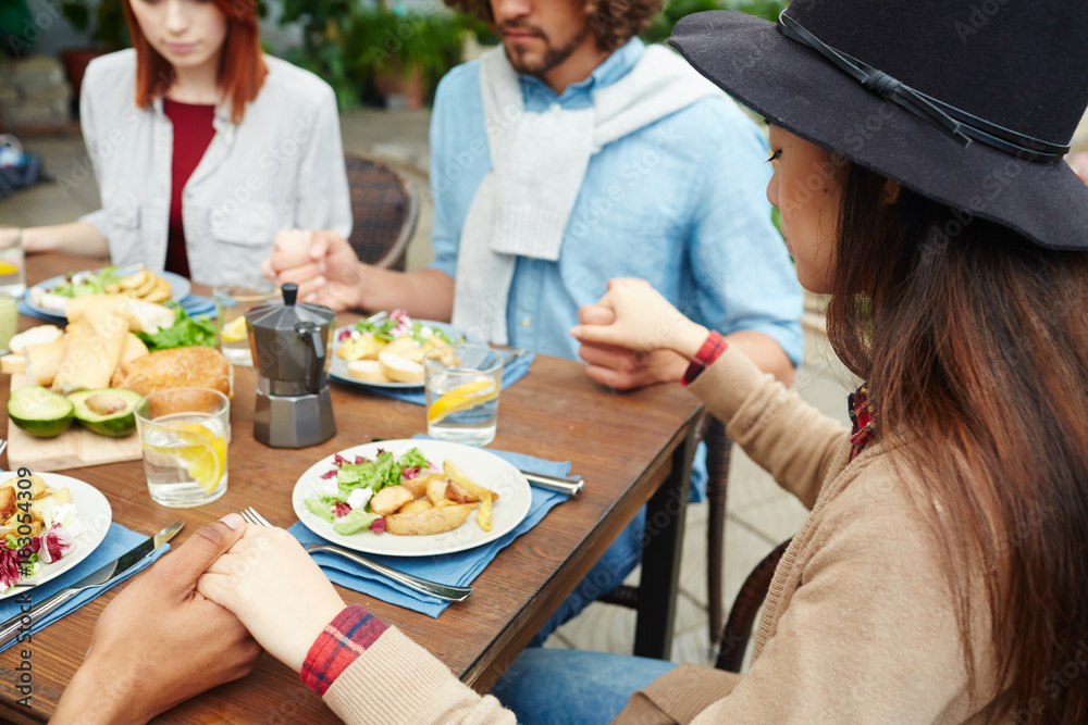 Company of friends holding by hands while sitting by served table and praying before dinner Company of friends holding by hands while sitting by served table and praying before dinner