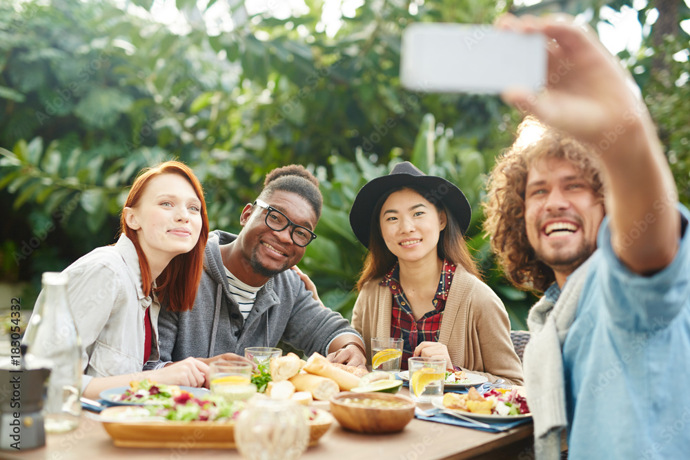 Group of international friends looking at smartphone camera while making selfie by served table