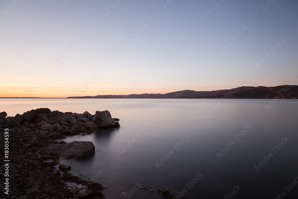 Dusk on a lake, with rocks in the foreground, perfectly still water and empty sky Dusk on a lake, with rocks in the foreground, perfectly still water and empty sky