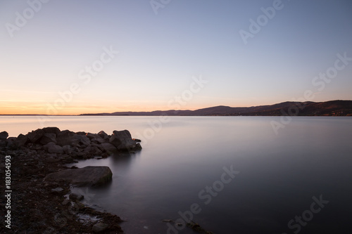 Dusk on a lake  with rocks in the foreground  perfectly still water  and empty sky