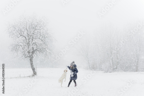 little kid, child playing with white dog on snow in winter day, flying snowflakes, trees and she wears winter clothes