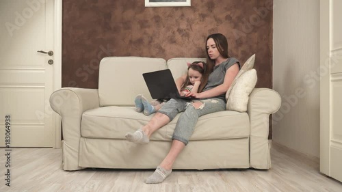 Mother and little daughter are sitting on the sofa and using laptop