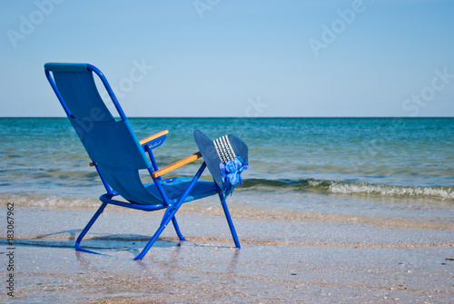 Fototapeta Naklejka Na Ścianę i Meble -  Blue beach chair and stylish blue hat made of straw with a large flower on the shore of the blue sea blue sky sea wave beautiful landscape summer vacation beach sand weekend rest shore