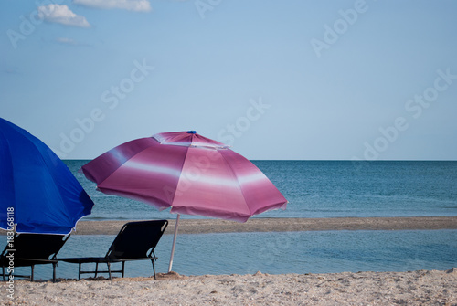 Two blue beach chairs and a blue and pink big umbrella for a shadow on the shore of the blue sea island of sand in the water blue sky white clouds sea wave beautiful landscape summer vacation beach © Elena