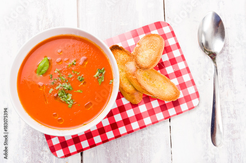 Fotografie Red tomato and bell pepper soup in a white bowl, served with toasted bread on a red and white checkered vintage cutting board on white wooden background