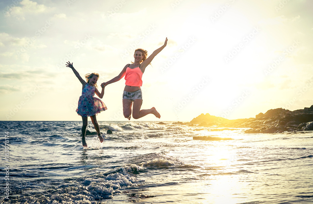 Fototapeta premium Szczęśliwa kochająca rodzina matki i córki, skoki w wodzie o zachodzie słońca na plaży - mama z dzieckiem, wspólna zabawa w letni dzień - koncepcja lato, podróże, wakacje i rodzina