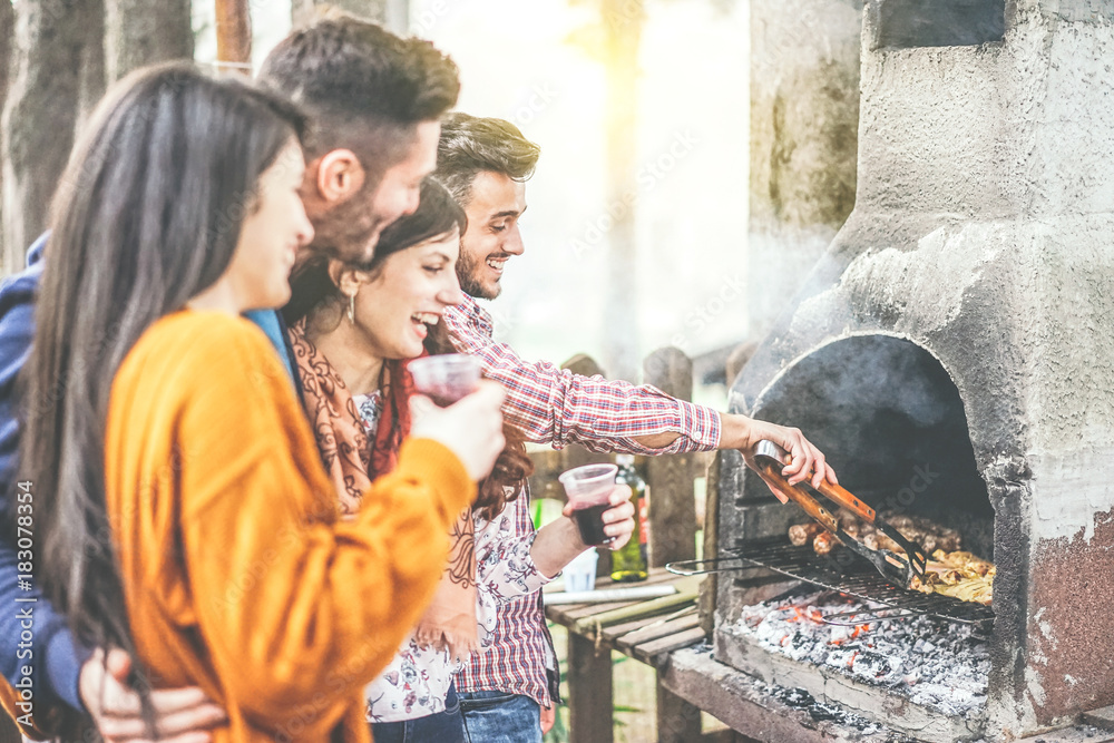 Young people having a barbecue party in the backyard on mountain - Happy friends grilling meat and drinking red wine outdoor - Friendship, lifestyle and food concept - Focus on male hand right