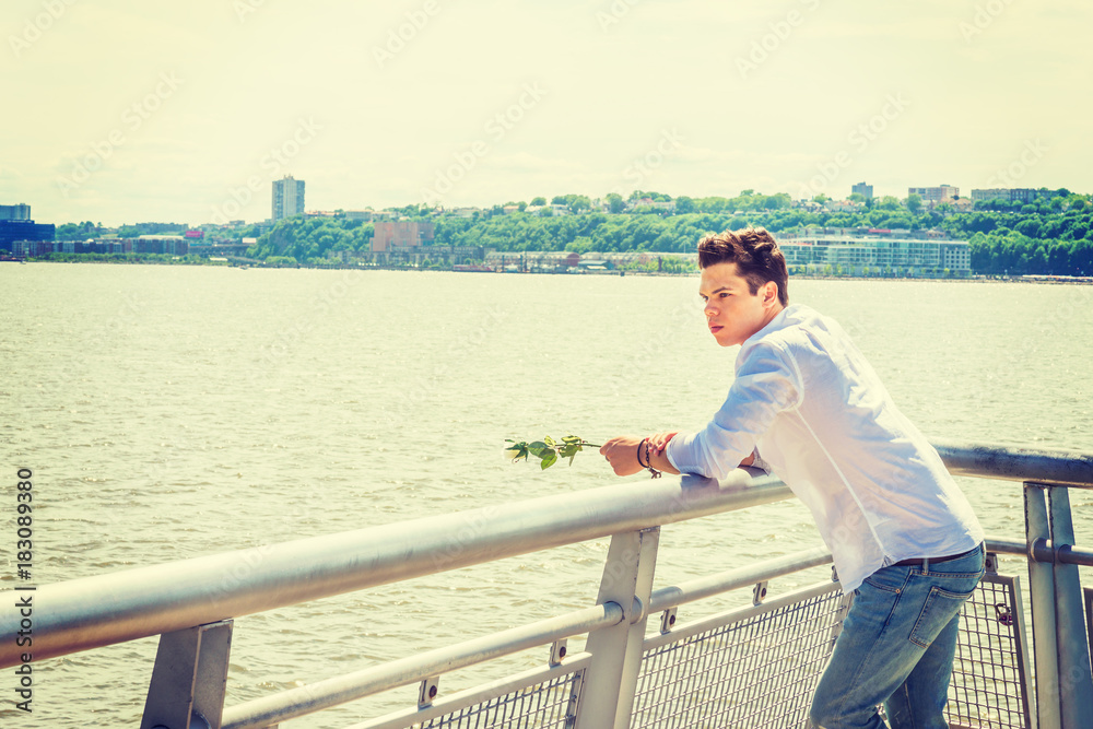 Man Missing You. Wearing white shirt, jeans, holding white rose, a guy standing by Hudson River in New York, opposite New Jersey, looking at white rose on hands, thinking, lost in thought.  Copy Space
