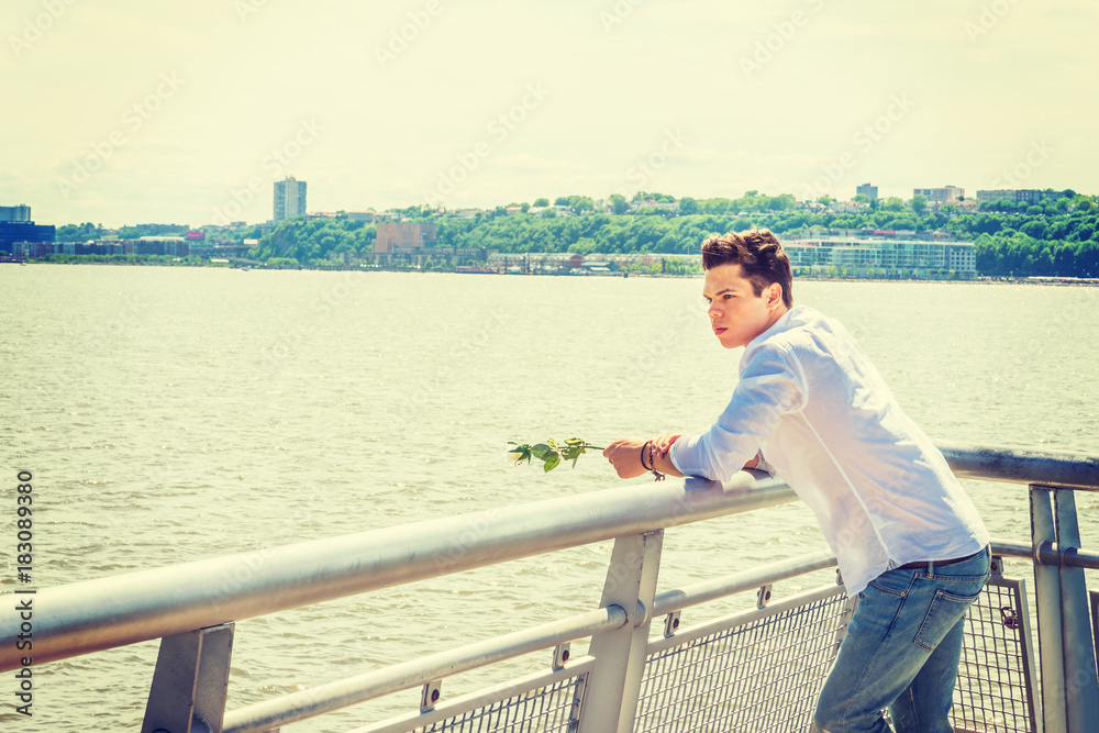 Man Missing You. Wearing white shirt, jeans, holding white rose, a guy standing by Hudson River in New York, opposite New Jersey, looking at white rose on hands, thinking, lost in thought. Copy Space Man Missing You. Wearing white shirt, jeans, holding white rose, a guy standing by Hudson River in New York, opposite New Jersey, looking at white rose on hands, thinking, lost in thought. Copy Space