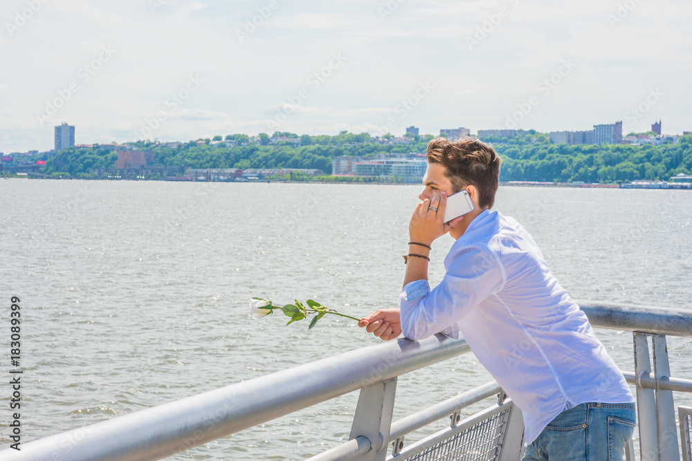 Wearing white shirt, jeans, holding white rose, a young guy standing by Hudson River in New York, opposite New Jersey, listening, talking on mobile phone. Concept of looking for love, friendship..