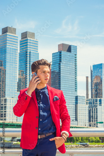 Businessman traveling, working in New York.Dressing in red blazer, blue collarless shirt, pants, a young guy standing outside busy business district with high buildings, talking on his mobile phone..