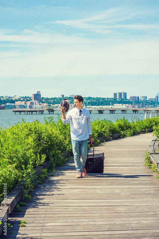 Man Traveling in New York. Wearing white shirt, jeans, sneakers, sunglasses hanged on collar, holding Fedora hat, dragging rolling luggage, a young guy walking on wooden road by Hudson River. 