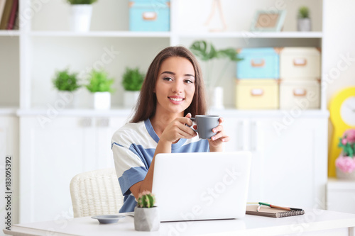Young woman using laptop and drinking coffee while sitting at table indoors