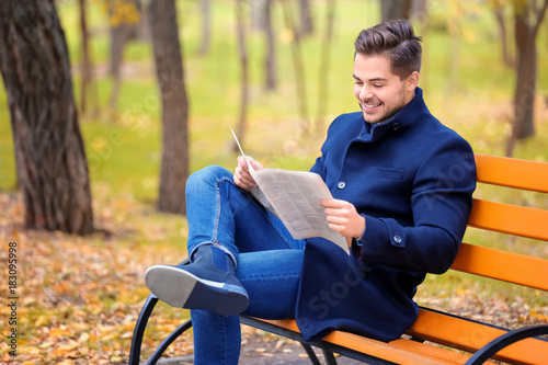 Young handsome man with newspaper sitting on bench in park