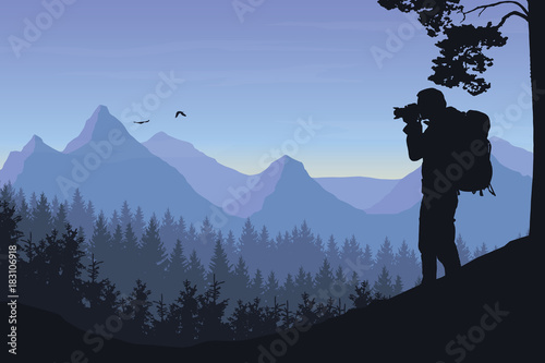 A tourist photographing a flying bird in a mountain landscape with forest under a morning sky with dawn and clouds