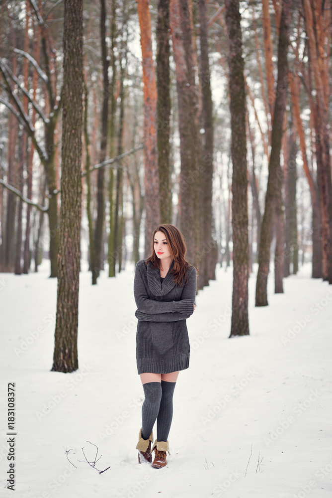 Winter portrait of a happy beautiful girl with brown hair in snow winter forest . Woman wearing knitted grey pullover dress and stockings high socks. Winter atmosphere, nature and freedom concept.