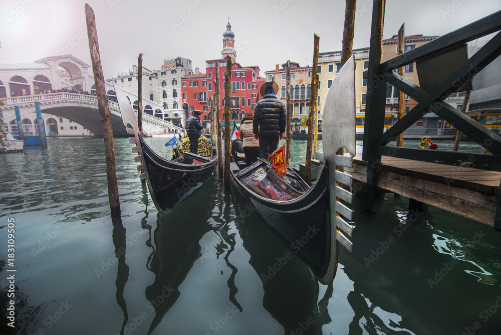 Gondoliers in gondolas at a canal in Venice with Rialto bridge in the background.