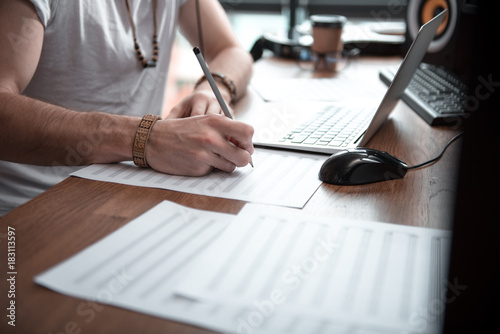 Close up of male hands drawing musical notes on paper. Man is sitting at desk near laptop