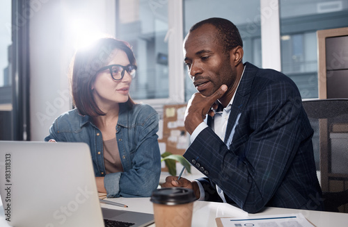 Need to think. Serious pleasant employees are sitting in office while discussing new project. Woman is looking at african man who is working on modern laptop with concentration and touching his chin