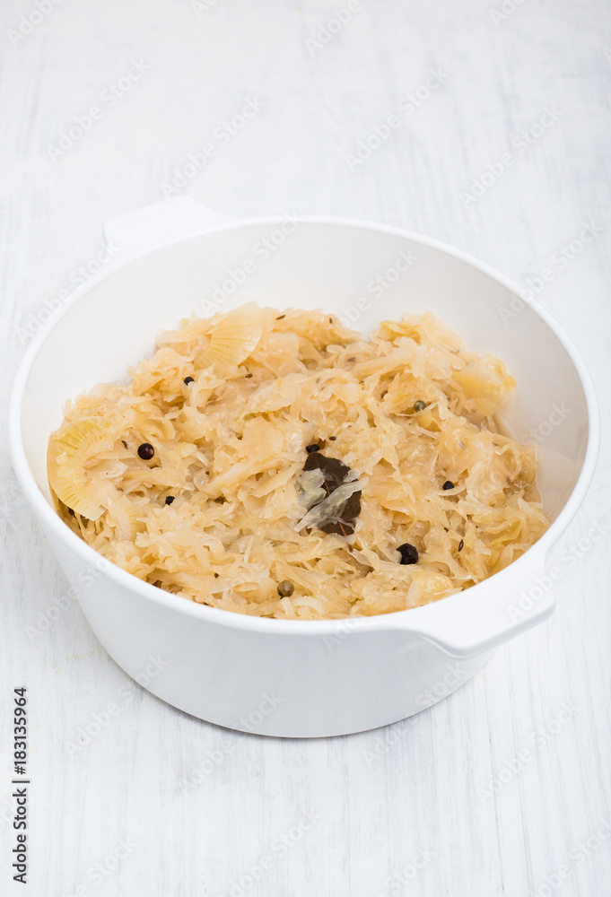 Traditional German Sauerkraut cooked with white wine, apples, onions, bay leaves, peppercorns, juniper berries, caraway and allspice in white pot on white wooden background Traditional German Sauerkraut cooked with white wine, apples, onions, bay leaves, peppercorns, juniper berries, caraway and allspice in white pot on white wooden background