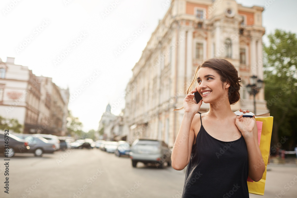 Portrait of young good-looking caucasian woman with dark hair in black dress waking around city, talking on phone, holding shopping bags in hands, relaxing after work. Lifestyle concept Portrait of young good-looking caucasian woman with dark hair in black dress waking around city, talking on phone, holding shopping bags in hands, relaxing after work. Lifestyle concept
