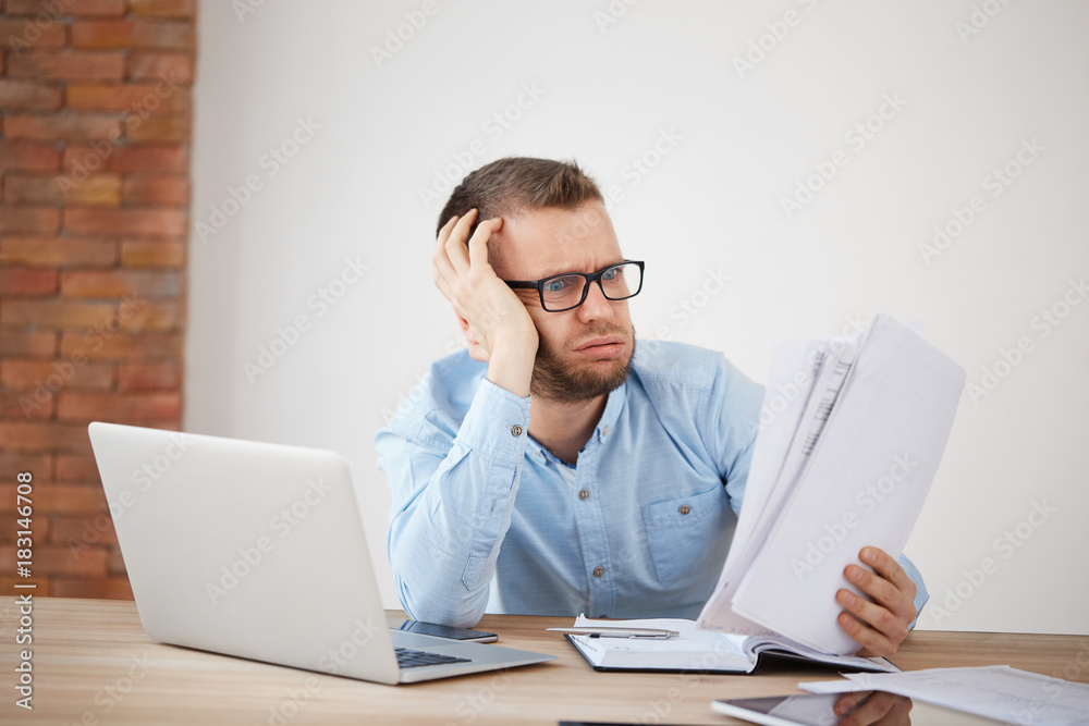 Close up of young exhausted unhappy bearded male finance manager sitting in office late in evening, being late home for family dinner, tired from big amount of work Close up of young exhausted unhappy bearded male finance manager sitting in office late in evening, being late home for family dinner, tired from big amount of work