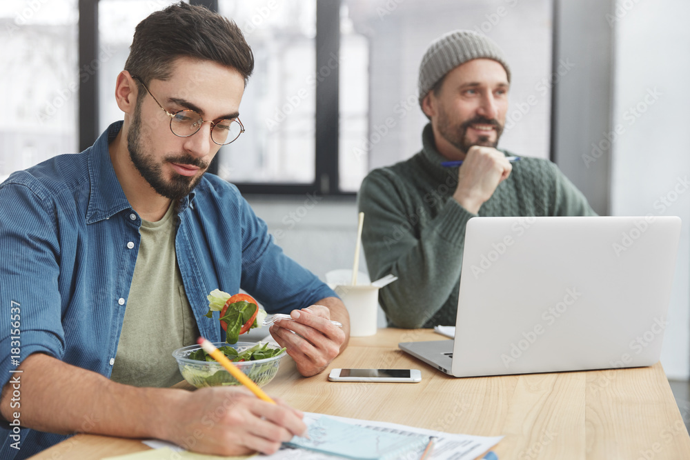Two coworkers works together at common project, dressed casually. Bearded young male works on documents, has fast snack, eats healthy salad and his partner in background uses laptop computer Two coworkers works together at common project, dressed casually. Bearded young male works on documents, has fast snack, eats healthy salad and his partner in background uses laptop computer