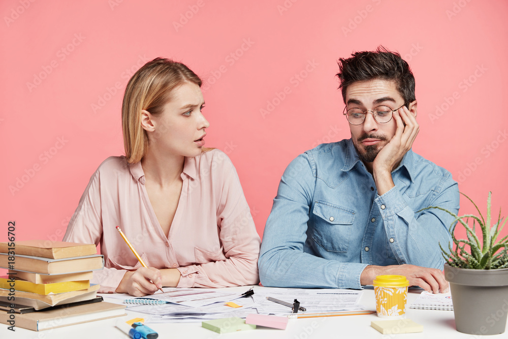 Young female and male students collaborate for making project, hipster guy look in bewilderment at notes, doesn`t know how to do, blonde woman tries make him working. People and work concept. Young female and male students collaborate for making project, hipster guy look in bewilderment at notes, doesn`t know how to do, blonde woman tries make him working. People and work concept.