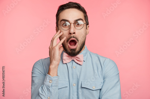 Shocked young handsome man stares through spectacles, being surprised to hear bad news or frightened of future difficuties on work, poses against pink background. Emotional bearded attractive guy © wayhome.studio 