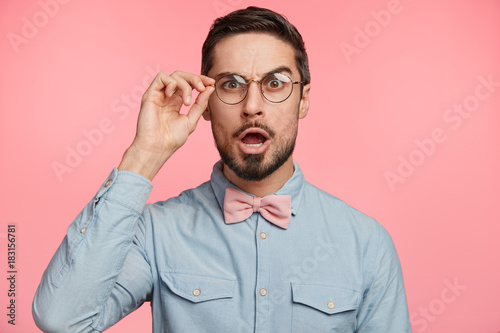 Surprised male manager stares through spectacles, keeps jaw dropped, shocked to see that there isn`t anyone on work place, tries to control work of colleagues. Amazed bearded young businessman