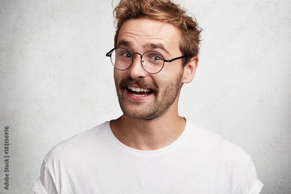 Portrait of cheerful smiling pleasant looking male guy wears round spectacles and casual t shirt, listens with great interest and pleasure interlocutor who tells interesting, intriguing, funny story Portrait of cheerful smiling pleasant looking male guy wears round spectacles and casual t shirt, listens with great interest and pleasure interlocutor who tells interesting, intriguing, funny story