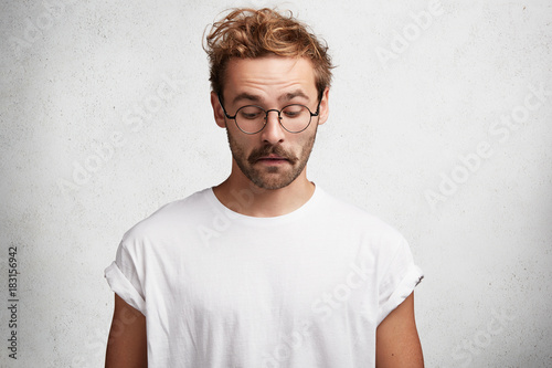 Curious intelligent male wears round spectacles, casual t shirt, notices something down, concentrated on floor, has puzzled look, isolated over white studio background. People and facial expressions