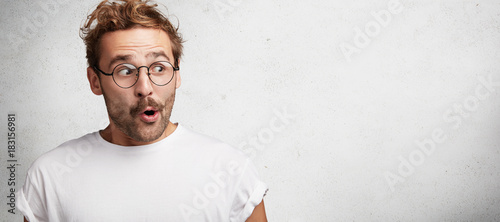 Horizontal shot of amazed bearded male has European appearance wears spectacles, looks in satisfaction aside, sees something wonderful and amazing, poses against white background with copy space