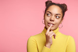Portrait of thoughtful African American female looks pensively aside with dreamful expression, notices someone, poses against pink background with copy space for your advertisment or hearder