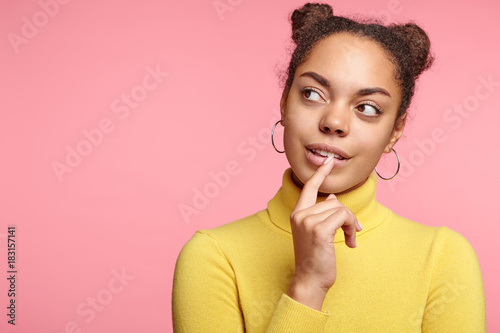 Portrait of thoughtful African American female looks pensively aside with dreamful expression, notices someone, poses against pink background with copy space for your advertisment or hearder
