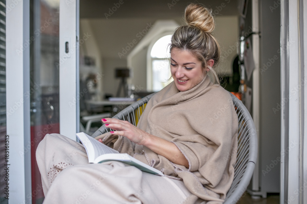 Young beautiful woman sits and reads a book on a terrace covered with a blanket and in the background there is a fireplace Young beautiful woman sits and reads a book on a terrace covered with a blanket and in the background there is a fireplace