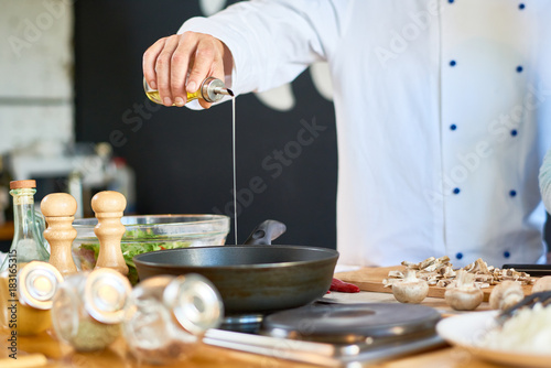 Close-up shot of unrecognizable chef wearing uniform pouring olive oil into frying pen while preparing mushrooms at modern restaurant kitchen