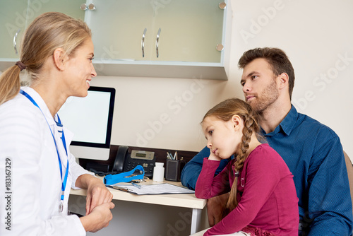 Portrait of smiling young pediatrician talking to man about health of his little girl, all sitting at desk in office